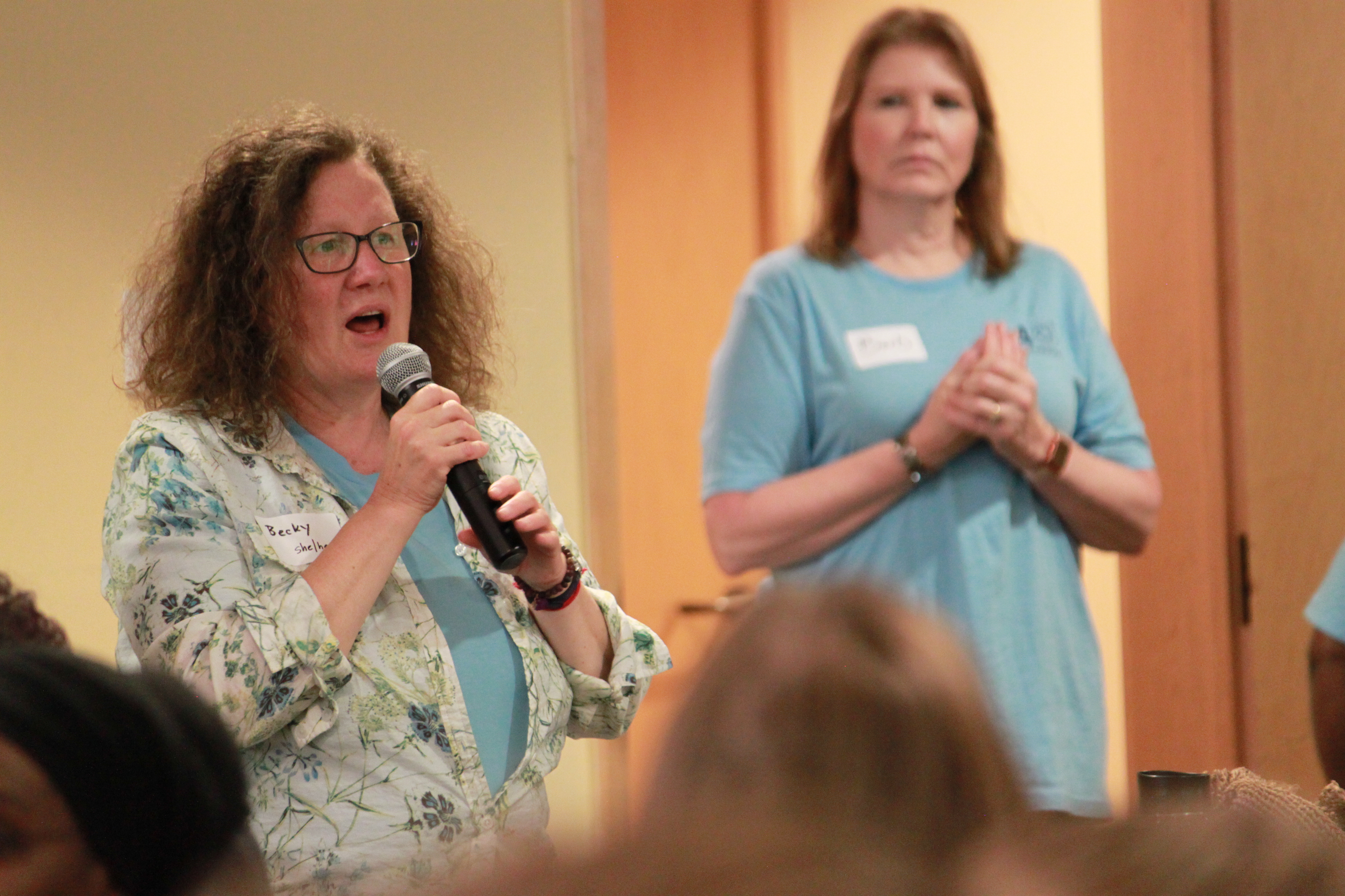 A woman holding a microphone while another woman listens in the background.